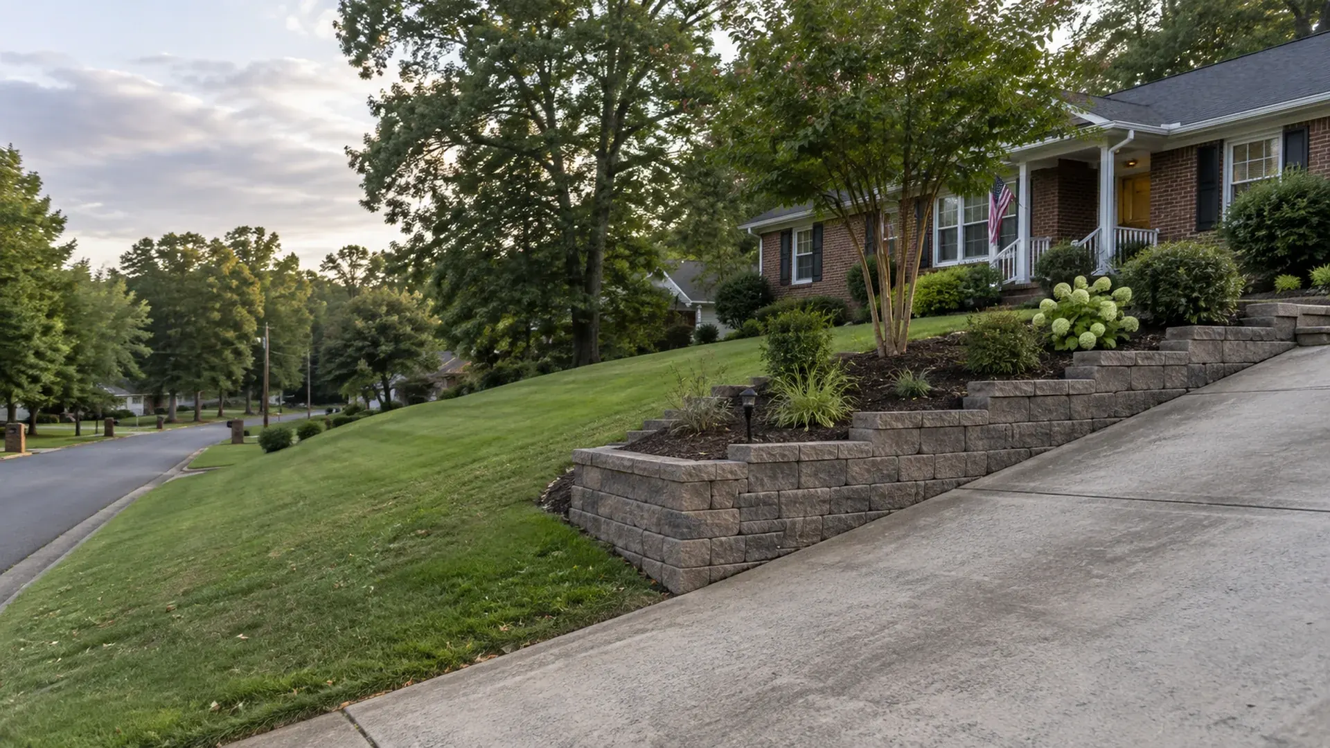 A sloped front yard along a residential road in the Chamberlayne area of Henrico, Virginia with a block retaining wall beside a driveway approach.