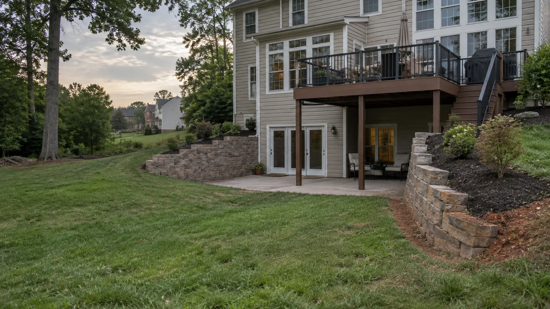 A walkout basement lot in a 1990s Midlothian, Virginia subdivision with segmental block retaining walls on both sides, red clay soil visible at the edge of fresh mulch.